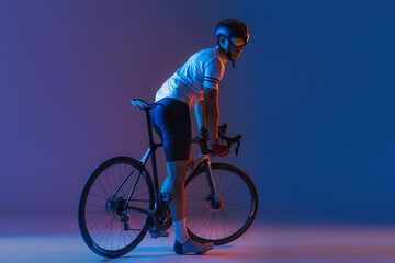 One male cyclist riding bicycle wearing cycling shorts and protective helmet isolated on dark blue background in neon. Concept of sport, speed, energy