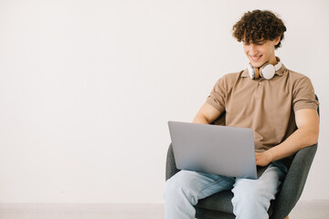 Attractive young man using laptop, wearing headphones while sitting in armchair against white wall, copy space. Millennial male communicating online, working or studying remotely on portable pc