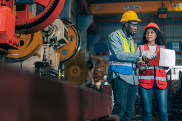 Two professional engineer/worker/technician use clipboard discuss work, walk in steel metal manufacture factory plant industry. Black African American man and woman wear hard hat check quality machine