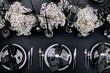 Top view of black style banquet table with a black tablecloth, silver plates, napkins, cutlery, black candles, and white flowers.
