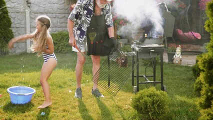 old man is grilling in the backyard while children are splashing with water balloons on a hot summer