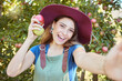 © Jade M/peopleimages.com - Selfie of a happy female farmer standing in an orchard holding two different apples. Portrait of one young smiling farm worker wearing a straw hat and dungaree on a sunny day picking fruit