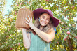Young smiling woman carrying a bucket filled with apples. One female holding a bag full of organic fruit in an orchard during harvest season outside. Farmer harvesting fruits from trees on a farm