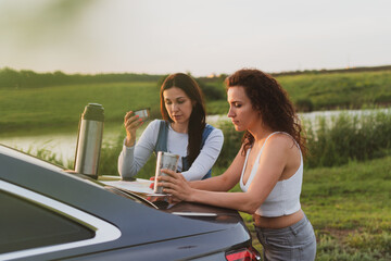 two girls are traveling on the roads by car, stopped at the side of the road and look at the map, drinking coffee from a thermos. Vacation concept