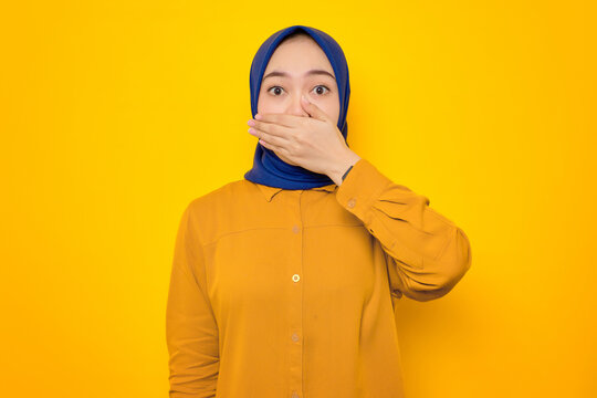 Shocked young Asian Muslim woman dressed in orange covering mouth with hand for mistake isolated on yellow background