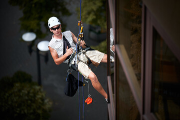 Wall Mural - Industrial mountaineering worker in protective helmet showing approval gesture thumbs up while hanging on climbing rope outside building. Man in sunglasses using safety lifting equipment outdoors.