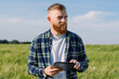 A male agronomist with a beard and a plaid shirt walks through a field with wheat with a tablet during an inspection. Against the backdrop of a megapolis