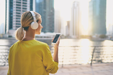 Wall Mural - Enjoying the sound. Happy young woman with earphones listening music on smartphone while walking on city embankment.