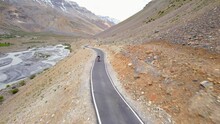 Aerial Drone Shot Of A Lone Female Or Woman Bike Rider Or Solo Biker With Saddlebag Traveling On Empty Scenic Road In Cold Arid Hilly Region Next To A River Valley With Snow Peaks In The Background. 