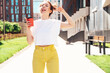 © halayalex - Young beautiful smiling hipster woman in trendy summer yellow jeans clothes. Carefree teen model posing in the street at sunset. Positive female drinks tea or coffee. Holding plastic cup