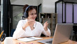 © wichayada - Smiling young asian woman in headset wave greet talking on webcam virtual conversation on laptop, happy female in wireless headphones speak on video call on computer, consult client online