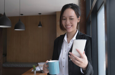 Happy young Asian businesswoman standing using smartphone and hold coffee cup  at office.