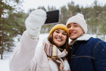 season, technology and leisure concept - happy couple with smartphone taking selfie in winter park