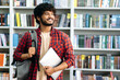 © Kateryna - Positive confident indian or arabian male student of university, in stylish casual wear, with backpack and laptop, stands in a library against the background of bookshelves, looks away, smiles, dreams