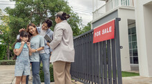 Female House Salesman, Home Broker, Standing In A Suit, Standing In Front Of A House For Sale Sign. There Are Family, Parents, Daughters Coming To Visit The Project Asking For Details Of The House For