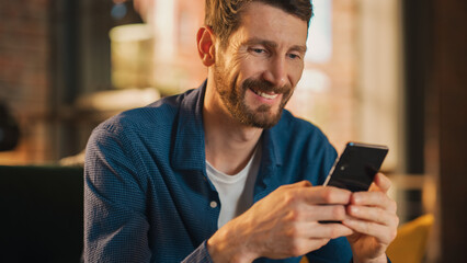 Poster - Portrait of a Handsome Man Sitting on Couch, Using Smartphone in Stylish Loft Apartment or Cafe. Creative Male Smiling, Checking Social Media and Typing Message. Urban City View from Big Window.
