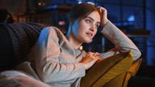 Portrait Of Beautiful Female Spending Time At Home, Resting On A Sofa, Watching TV Show Or News Broadcast In Stylish Loft Apartment. Young Woman Watching Movie Streaming Service Without Emotions.