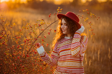 Happy smiling fashionable curly brunette woman wearing trendy autumn patterned sweater, stylish hat, posing in autumn nature during sunset. Fashion, lifestyle conception. Copy, empty space for text
