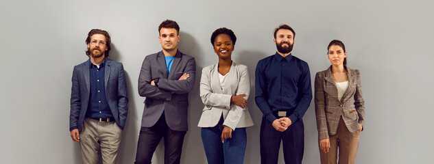 Confident business team in modern smart clothes. Group of happy young diverse people in formal suits standing in fashion studio and leaning on gray wall. Office dress code concept. Banner, header