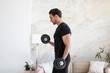 © Vitaliy - Side view of young handsome man in black sportswear doing biceps curl with dumbbells in living room at home. Healthy lifestyle and home workout concept