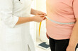 Nutritionist measuring overweight woman's waist with tape in clinic, closeup