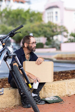 Homeless Man With Bicycle Sitting On Low Wall With Cardboard Sign