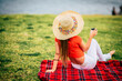 © simona - Back view of trendy female tourist relaxing sitting on a red cover in a meadow field wearing straw hat enjoying the view and the silence. Concept of alone leisure serene outdoors activity at the park