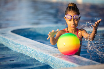 Wall Mural - A girl in a bright bathing suit swims with an inflatable ball in a pool with clear water on a summer evening