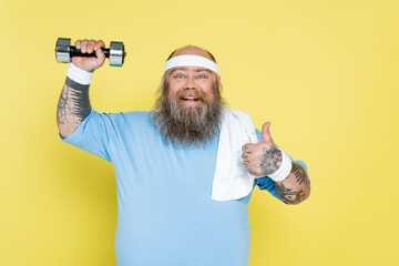 Wall Mural - cheerful plus size man showing thumb up while exercising with dumbbell isolated on yellow.