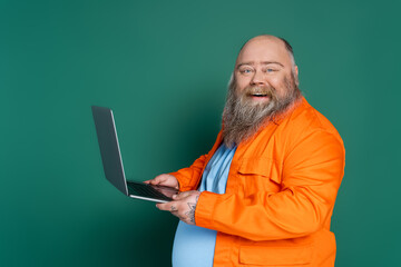 happy plus size man in orange shirt holding laptop and looking at camera isolated on green.