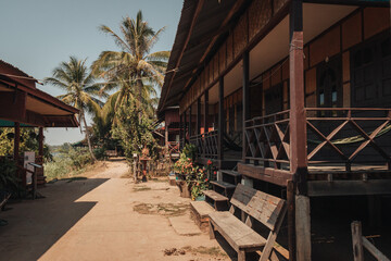 Wooden houses on stilts on a street on Don Det Island, Laos with palm trees in the background on a sunny day.