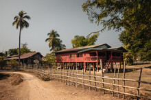 Don Det, Laos - January 18th, 2020 : Colorful Red And Green Lao Wooden House On Stilts With Palm Trees In The Background And A Bamboo Fence And Sandy Pathway In The Foreground.