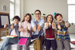 © Studio Romantic - Happy teacher with pupils group giving thumbs up. Toothy smiling tutor with elementary or secondary school student looking at camera portrait. Schoolroom interior. Offline education acknowledgment