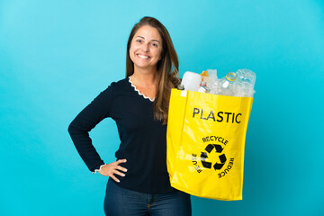 Wall Mural - Middle age Brazilian woman holding a bag full of plastic bottles to recycle on blue background posing with arms at hip and smiling