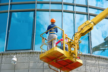 Female Window Cleaner Cleaning Glass Windows On Building At Height In Lifting Platform. Worker Polishing Glass, Window Washing On Office Building In Crane Bucket. Window Cleaners Exterior Glass Facade