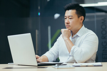 Wall Mural - Pensive and serious asian businessman working in office, thinking over projector, working with laptop.