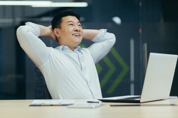 Dreamy successful Asian businessman working in office, smiling, hands behind head, thinking and building new dreams and plans.