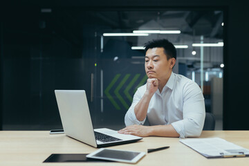 Wall Mural - Pensive asian businessman working in modern office, man thinking about decision, experienced director with laptop.