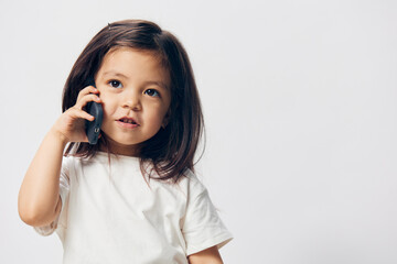 Wall Mural - a little girl of preschool age, sitting on a white background in a white T-shirt, enthusiastically talking on the phone, turning half-side to the camera. The topic of the conversation on the phone