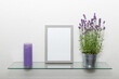 Blank grey wooden photo frame, lavender flowers in zinс pot, violet vintage candle on glass shelf against light wall.