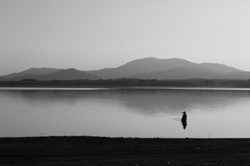 Wall Mural - A man is fishing in the lake at sunset. Wetland and mountain landscape. Black and white.