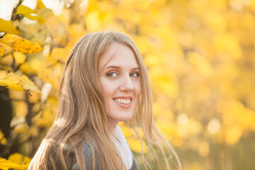 Wall Mural - Portrait of a beautiful young woman against a background of colorful golden foliage in an autumn park. Attractive woman with light brown hair near bushes with yellow leaves. Fall season. Close-up.