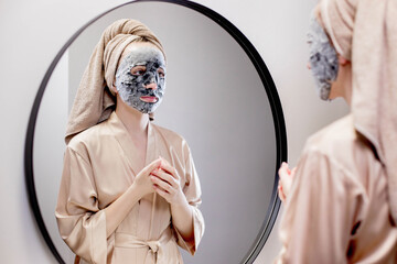 Wall Mural - Young woman applying face mask in bathroom and smiling. Beautiful woman in front of a mirror doing beauty treatments.