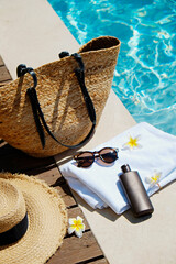 Swimming pool essentials concept. Beach bag with items for safe sunbathing on the deck, sunglasses, straw hat, white blanket & sunscreen product. Flat lay, copy space, top view, background.