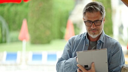Middle age man using tablet computer outdoor in garden, serious, concentrating. Home or holiday resort hotel. Browsing internet, online work, home office .