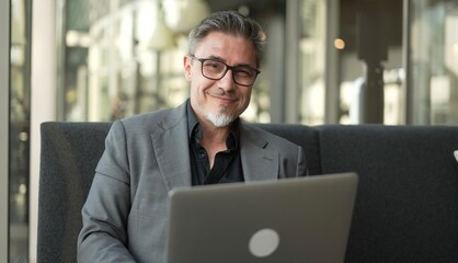 Canvas Print - Businessman sitting on couch working with laptop computer in office lobby. Portrait of happy middle aged man in business casual, smiling.