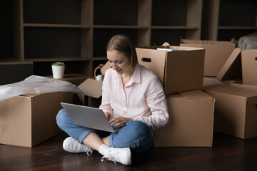 Wall Mural - Focused positive renter woman typing on laptop on moving, using computer at heap of boxes on floor in empty apartment, searching web for shipping, packing, transportation service