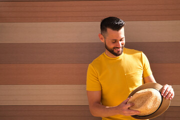 A young, handsome, blue-eyed, bearded man with a yellow T-shirt holds a hat in his hands. In the background a wall in shades of brown. The man smiles happily looking forward to the holidays.