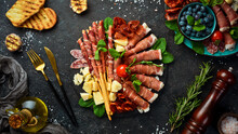 Italian Ingredients: Ham, Prosciutto, Salami, Parmesan, Olives, Bread Sticks. On A Black Stone Background.