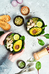 Wall Mural - Female hands holding a bowl of vegan lunch: avocado, tofu cheese, flax, pumpkin seeds and spinach. The concept of healthy eating. Top view.